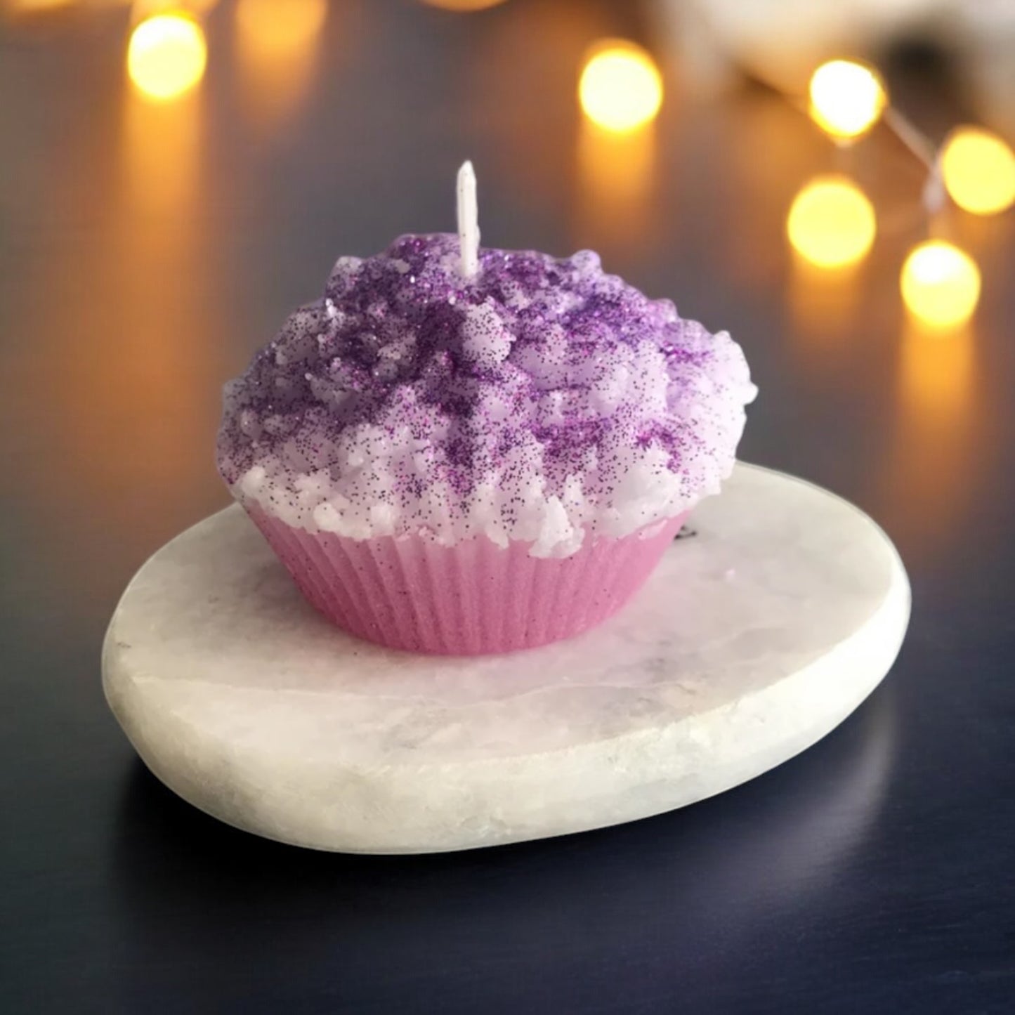 Detailreiche Muffin-Kerze mit lavendel-weißem Wachs-Topping, kunstvoll arrangiert auf einer Steinplatte mit warmen Lichtern im Hintergrund.